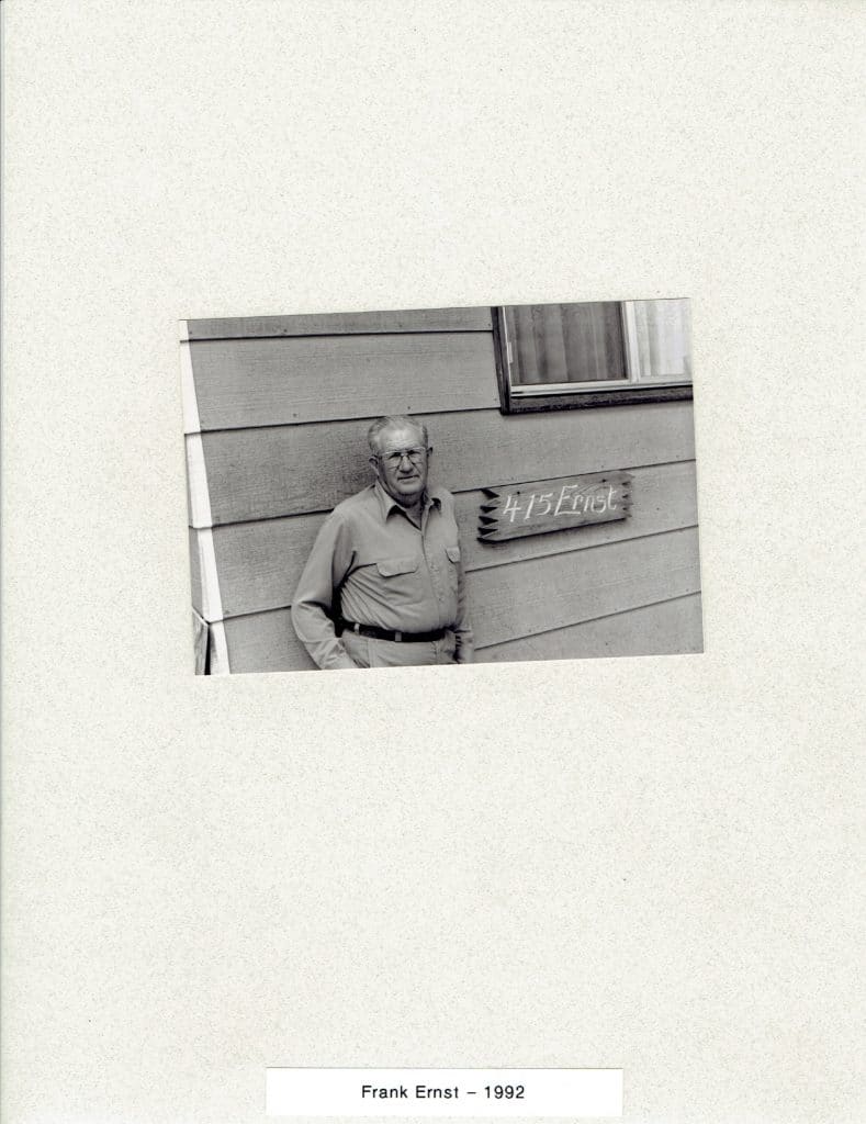 A black and white photo shows an older man in glasses and a shirt, standing by a house with "415 Ernst" on a sign.