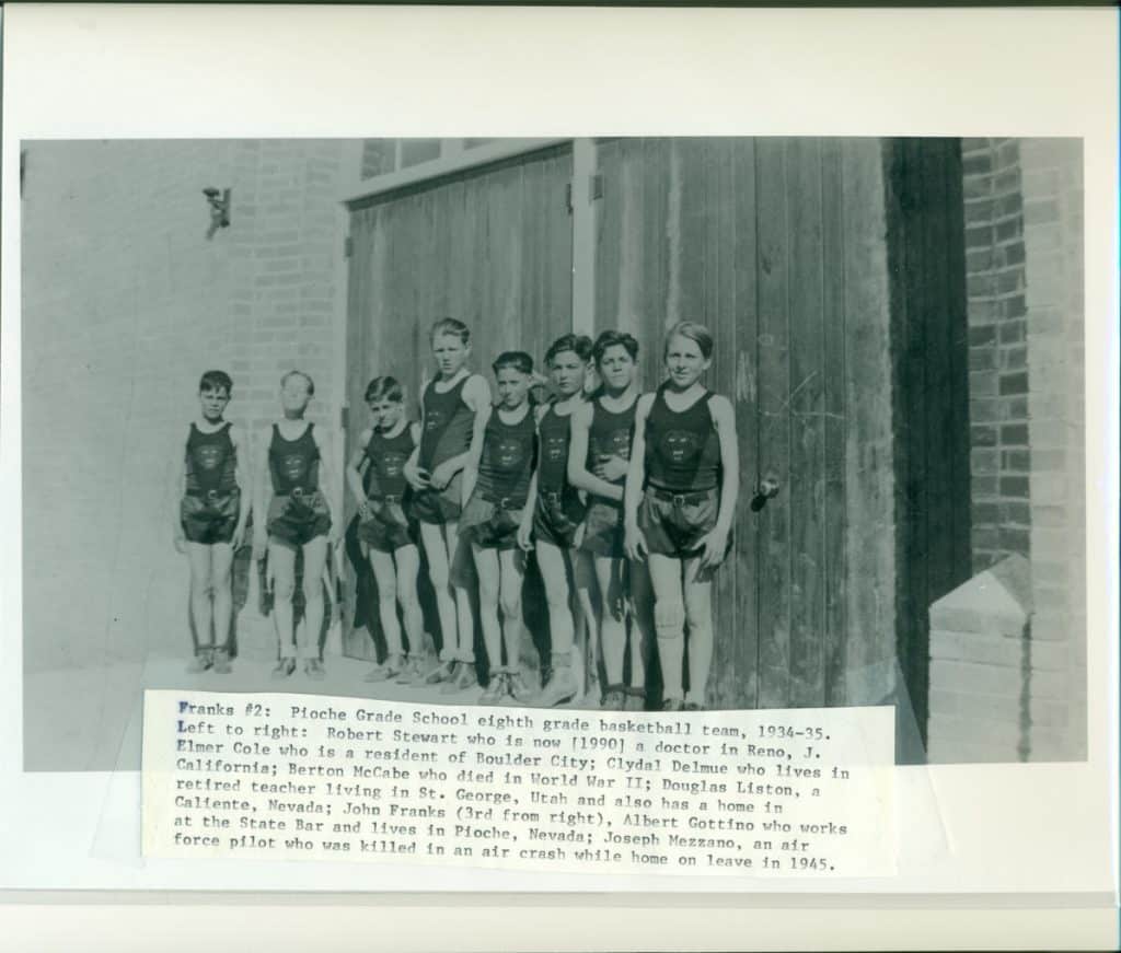 Nine young boys in basketball uniforms stand in front of a wooden door and brick wall.
