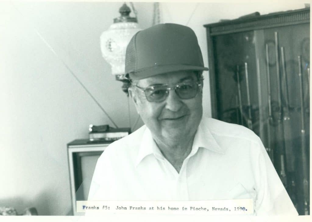 A man in a baseball cap and glasses smiles, with a decorative lamp and gun cabinet behind him.