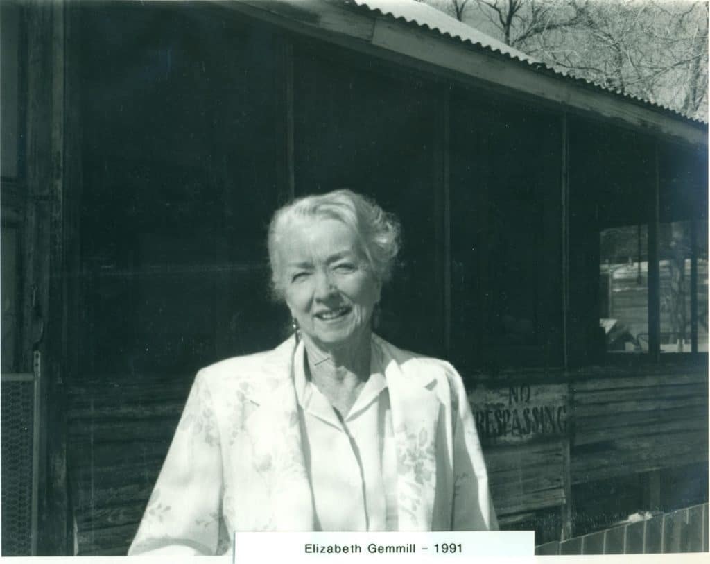 A smiling older woman with gray hair and a white jacket stands in front of a wooden building with a "NO TRESPASSING" sign.