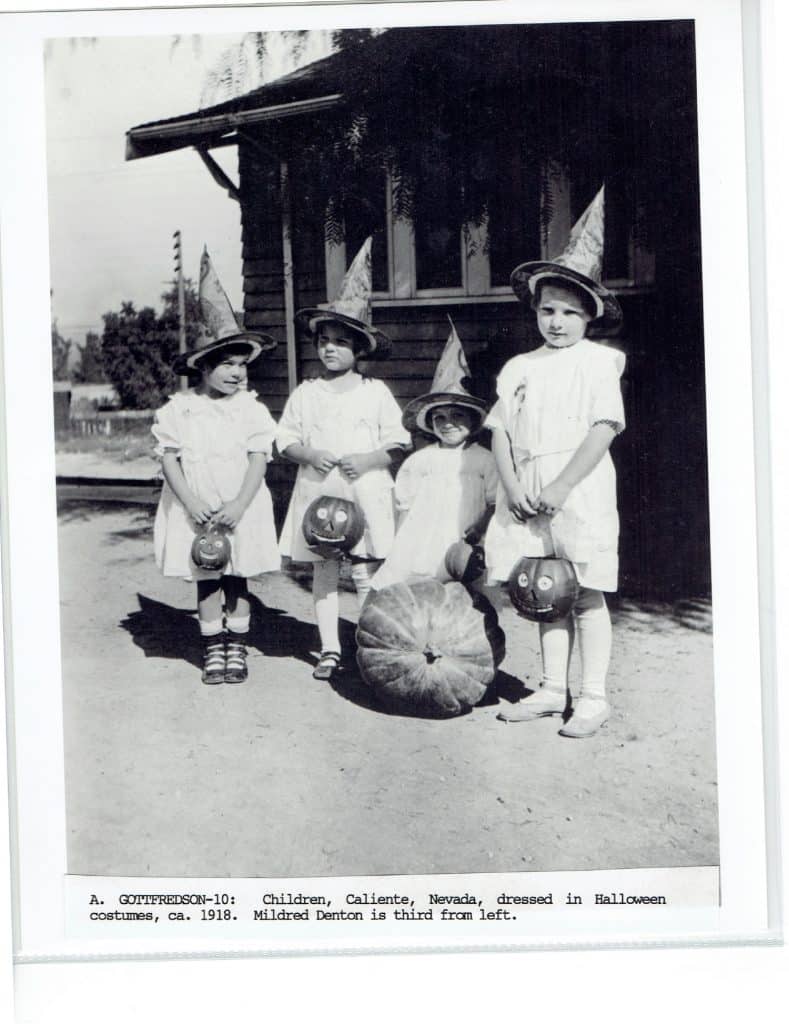 Four children in white dresses and pointed hats hold jack-o'-lantern buckets, with a pumpkin at their feet.