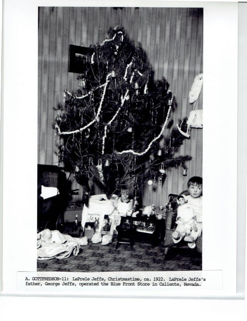 A black and white photo of a decorated Christmas tree with a child holding a doll in a chair.