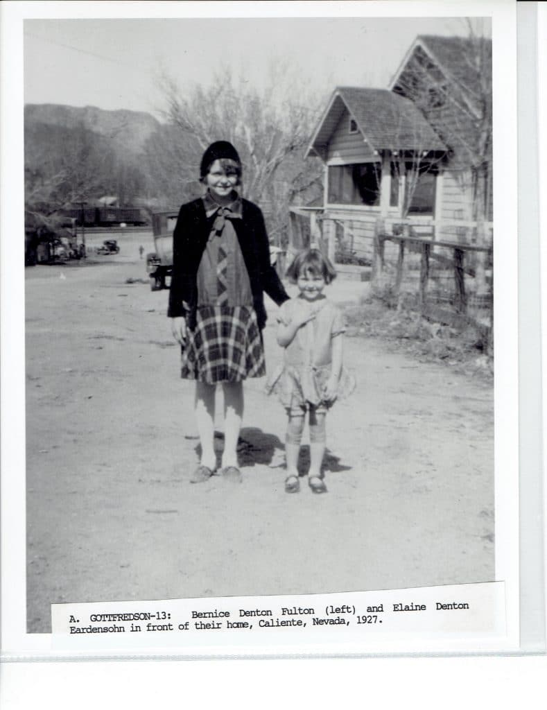 Two smiling girls stand on a dirt road in front of a house, with mountains in the distance.