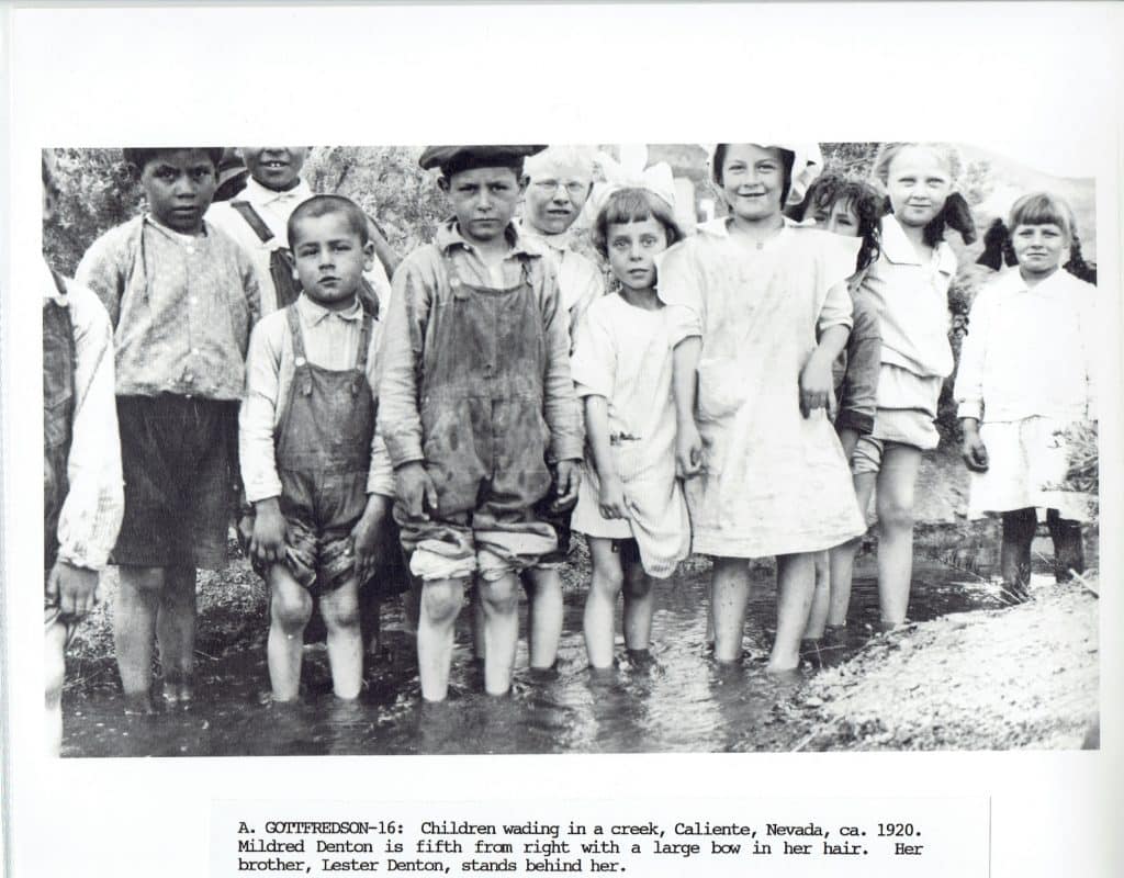 A group of children, some smiling, stand barefoot in a shallow creek, with trees in the background.