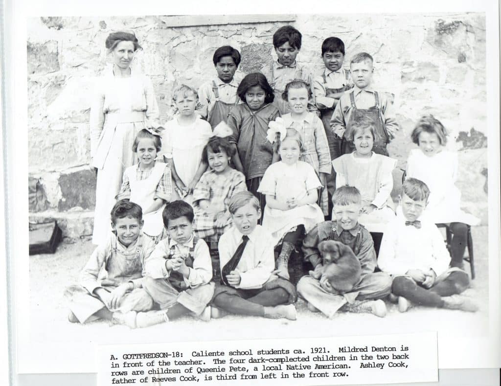 A black and white photo shows a female teacher and 20 children, some holding a puppy, posing outdoors.