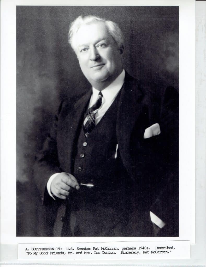 A black and white portrait shows a smiling man with white hair in a suit, holding glasses.