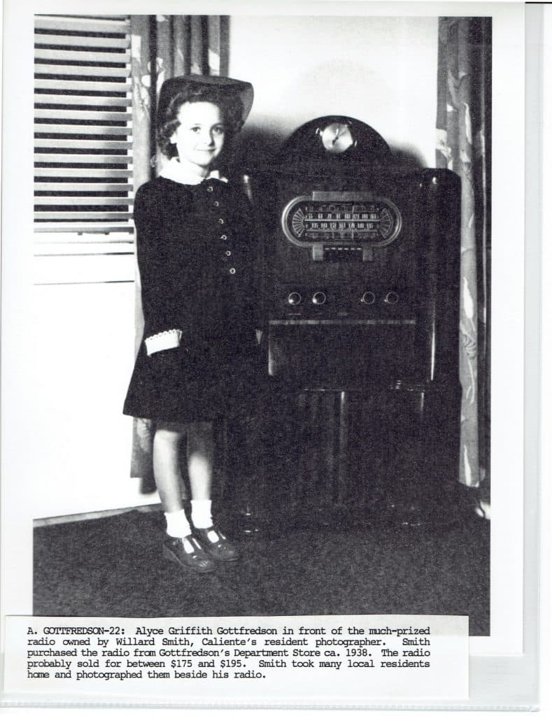 A young girl in a dark dress and hat stands smiling next to a large, ornate radio.
