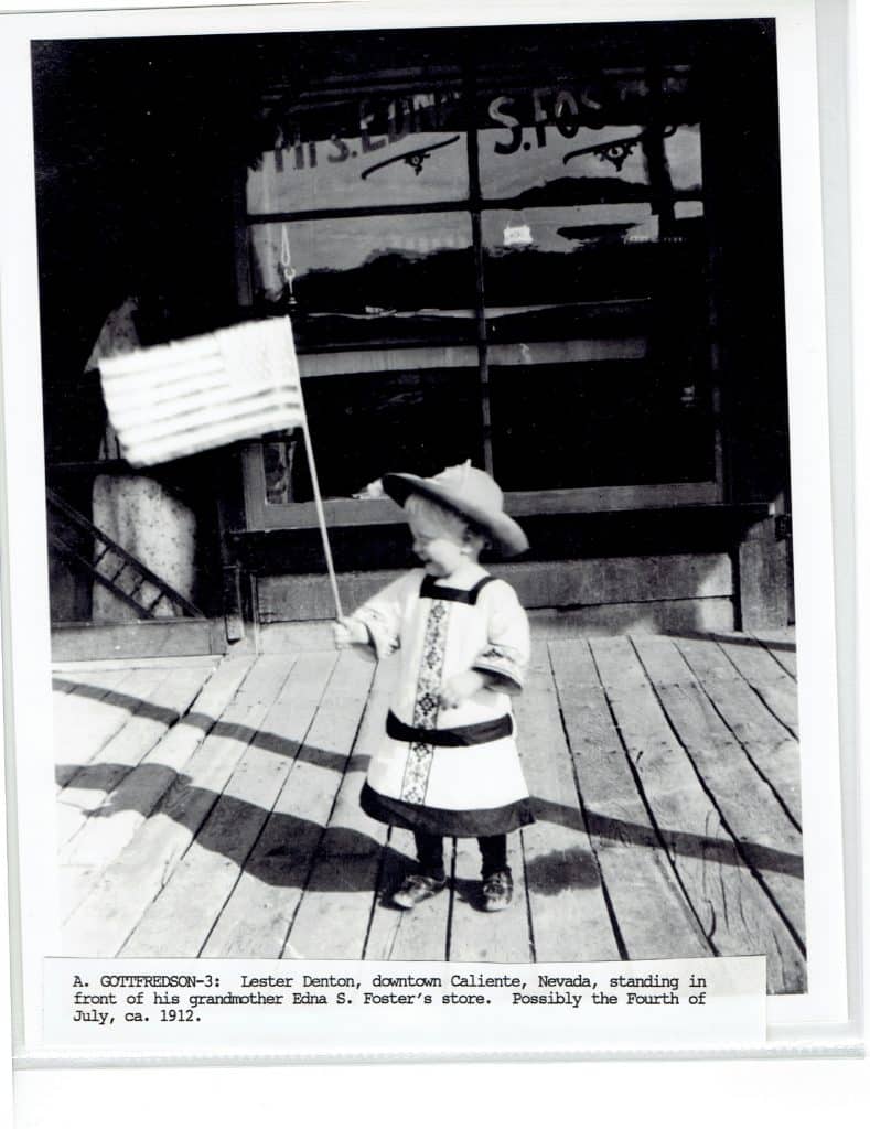 A toddler in a hat and dress holds an American flag on a wooden porch in front of a store window with "S. FOSTER" visible.