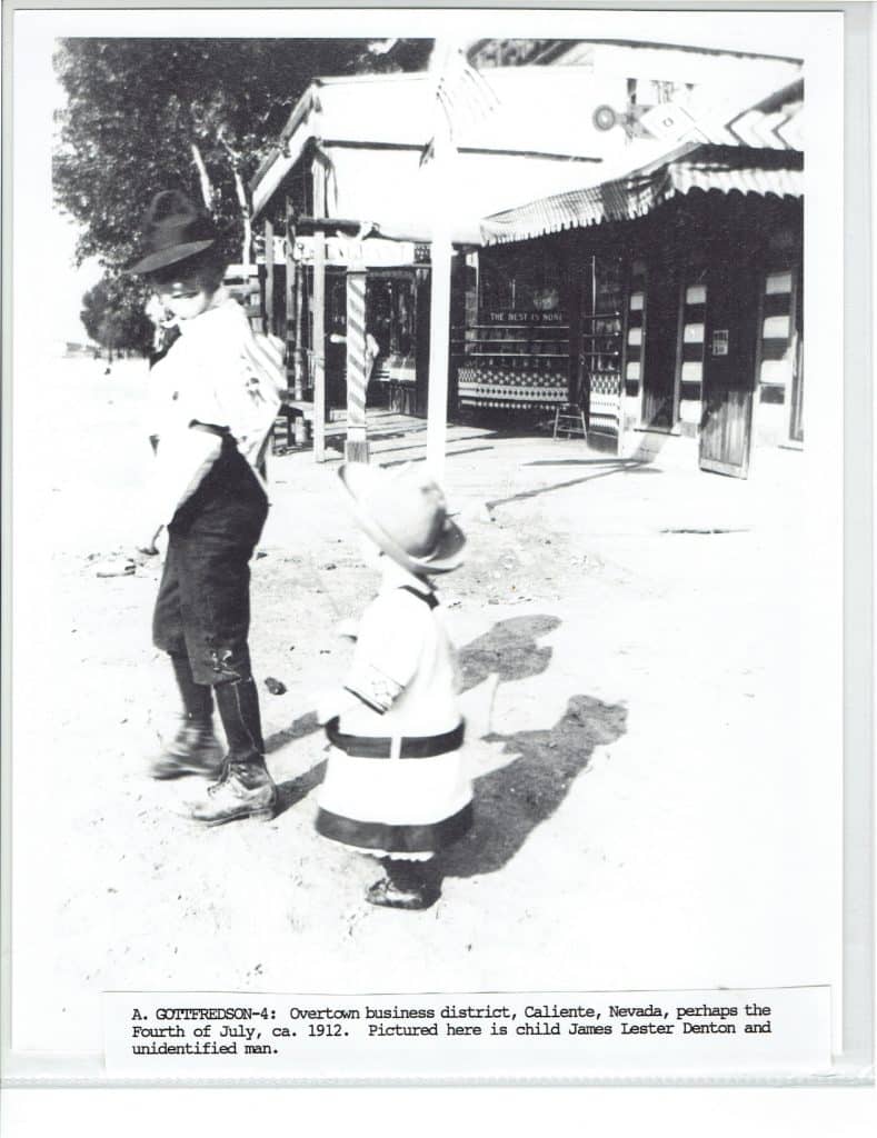 A black and white photo shows a man and a small child in hats standing on a dirt street.