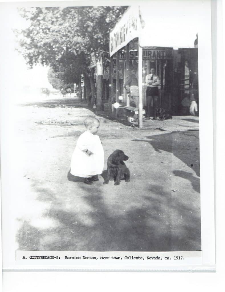 A black and white photo shows a toddler in a white dress standing next to a black dog on a dirt road.