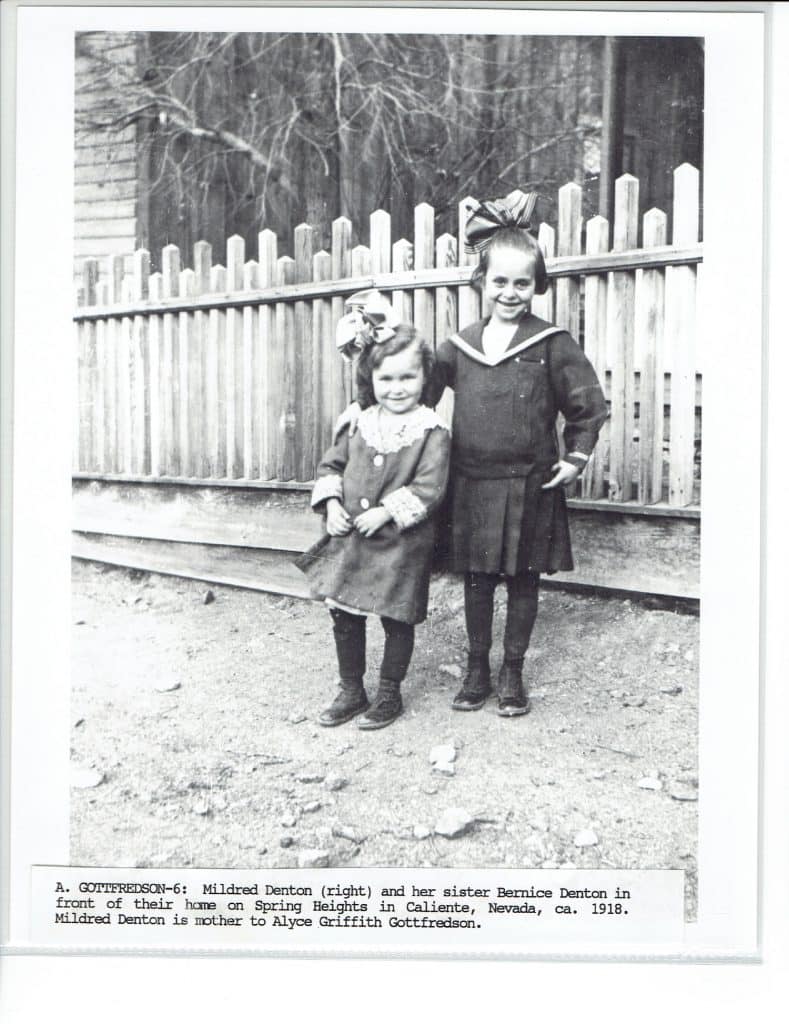 Two young girls in dark dresses and bows stand smiling in front of a picket fence.