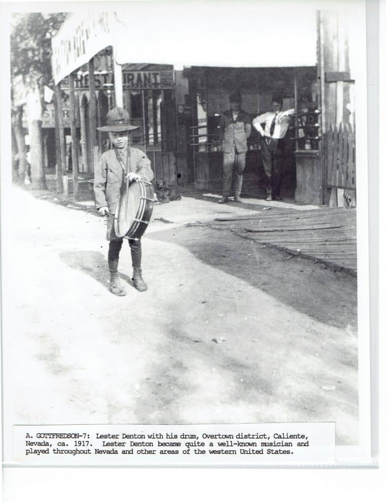 A young boy in a hat holds a drum on a dirt street in front of a building with "RESTAURANT" visible.