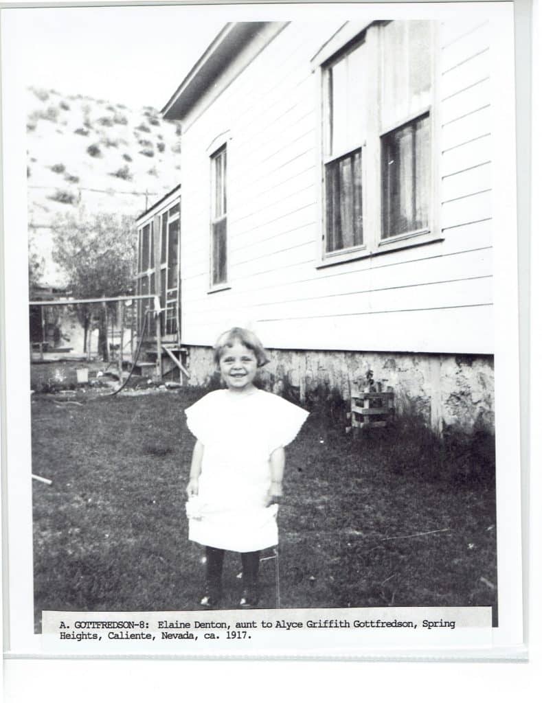 A smiling young girl in a white dress stands in a grassy yard in front of a house.