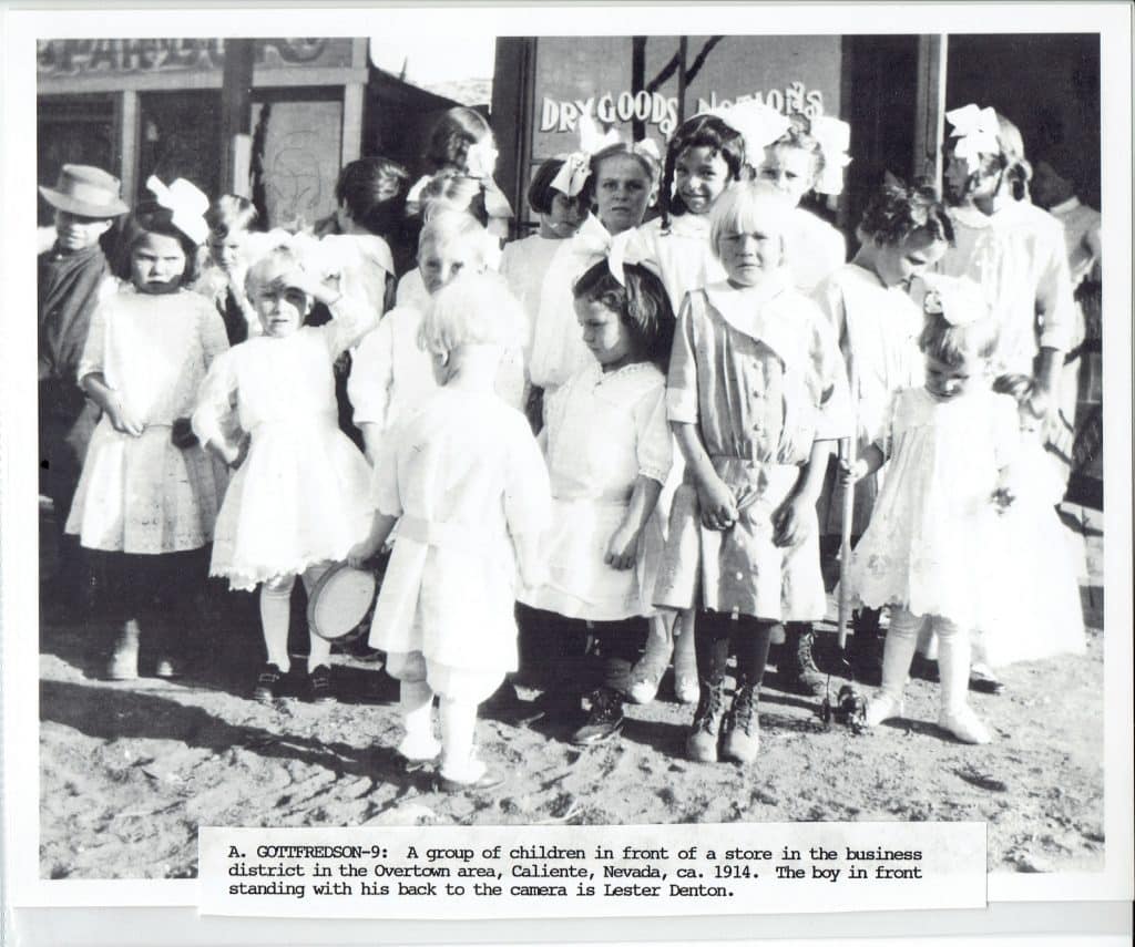A group of children, mostly girls in white dresses, stand outside a store with a "DRY GOODS" sign.