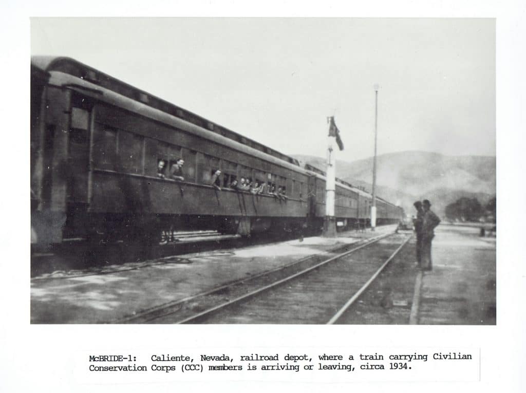 A black and white photo shows a train with passengers looking out windows, two men stand on the tracks.