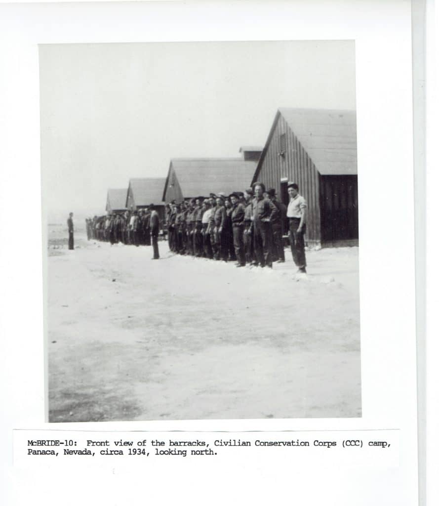 A black and white photo shows men in uniform standing in rows before several wooden barracks.