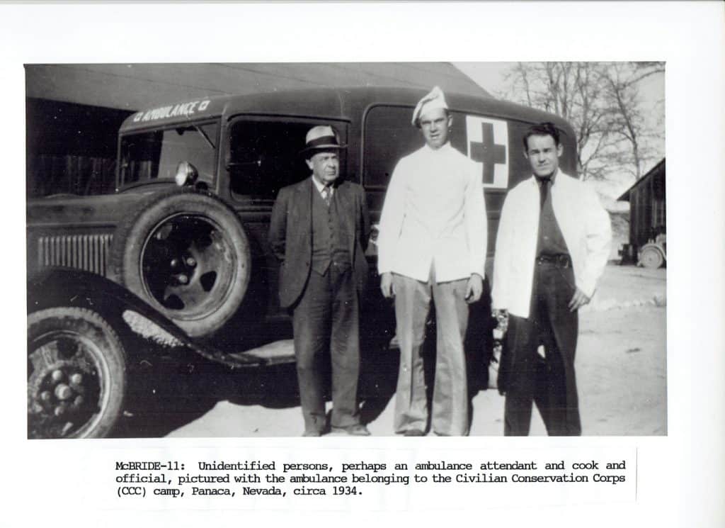 Three men stand in front of a 1930s ambulance with "AMBULANCE" and a Red Cross symbol on its side.