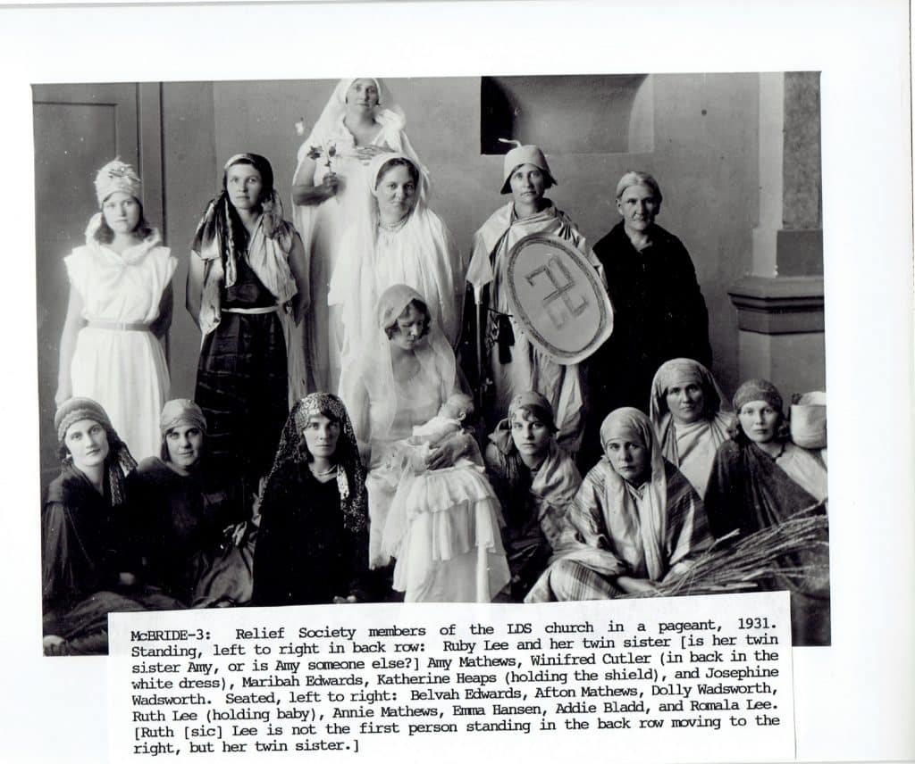 A black and white photo shows 15 women in costumes, some standing and some seated, with a swastika symbol on a shield.