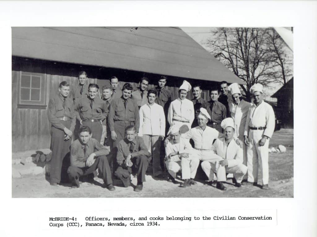 A black and white photo shows a group of 20 men, some in military uniforms and some in chef's whites, posing outside a wooden building.