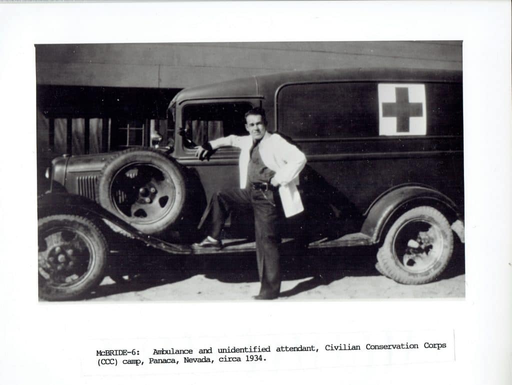 A man in a white coat stands with one foot on the running board of a vintage ambulance with a red cross.