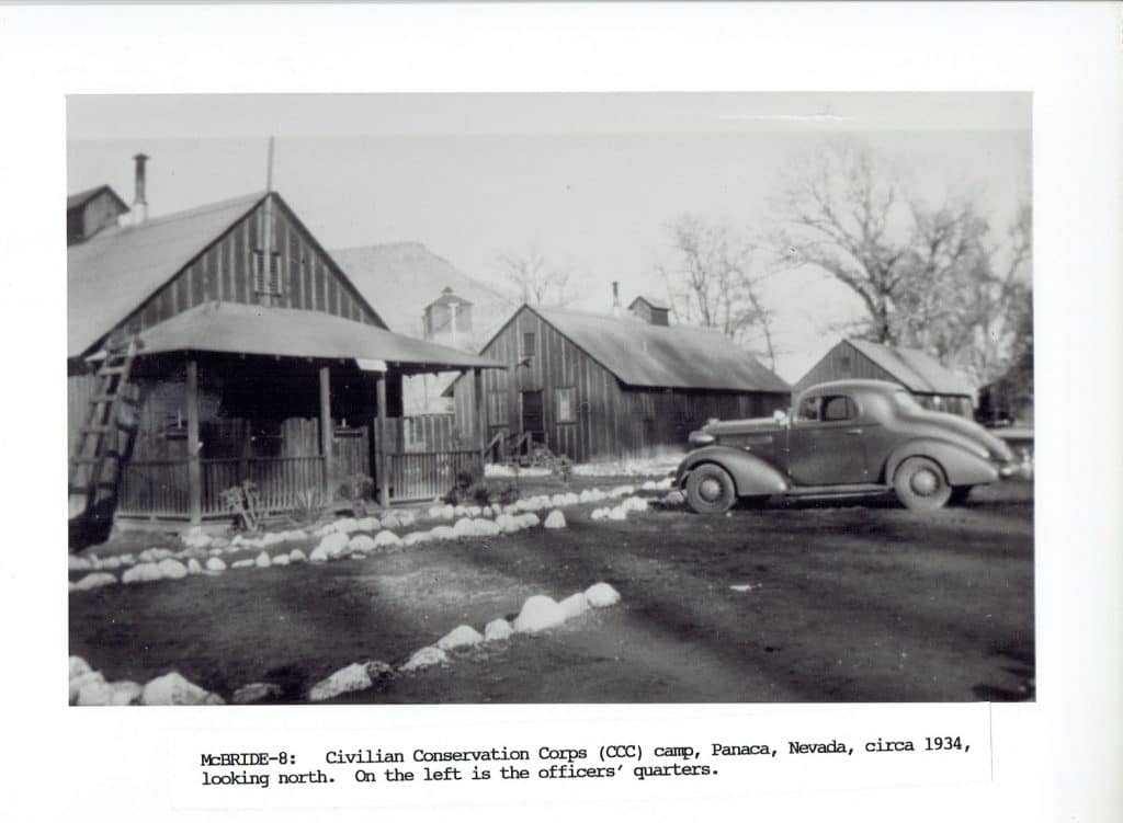A black and white photo of a 1930s car parked in front of wooden buildings at a CCC camp.
