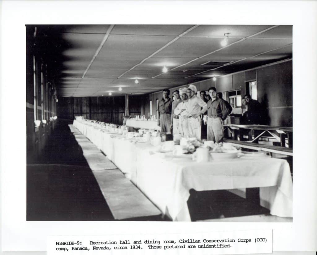 A long, narrow dining hall with multiple tables set for a meal and several men standing at the far end.