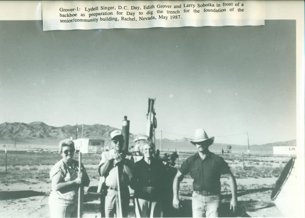 Four people stand in a desert landscape with a backhoe, mountains, and "Grover-1: Lydell Singer, D.C. Day, Edith Grover and Larry Sobotka in front of a backhoe as preparation for Day to dig the trench for the foundation of the senior/community bui...