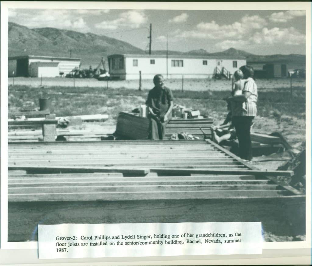 Two women and a child stand on a partially constructed wooden floor with trailer homes and mountains in the background.