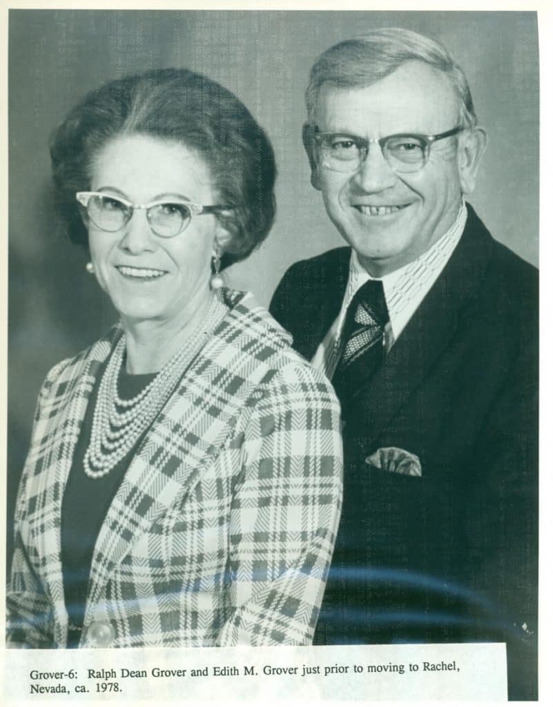 A smiling woman in a plaid jacket and pearl necklace stands next to a man in a suit and tie.