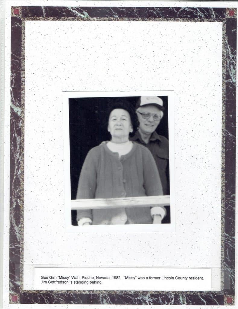 A black and white photo of an older woman in a cardigan and a man in a hat behind her, with text below: "Gue Gim "Missy" Wah, Pioche, Nevada, 1982. "Missy" was a former Lincoln County resident. Jim Gottfredson is standing behind."