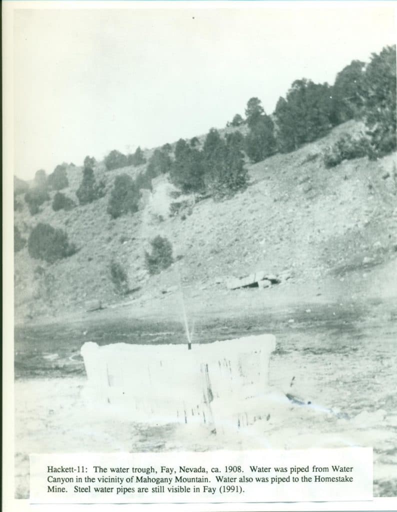 A black and white photo shows a water trough with ice formations, spraying water, against a barren hillside.