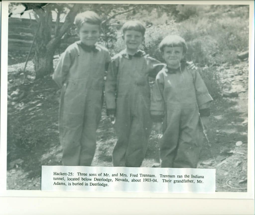 Three smiling boys in light-colored jumpsuits stand outdoors, with a tree and brush behind them.