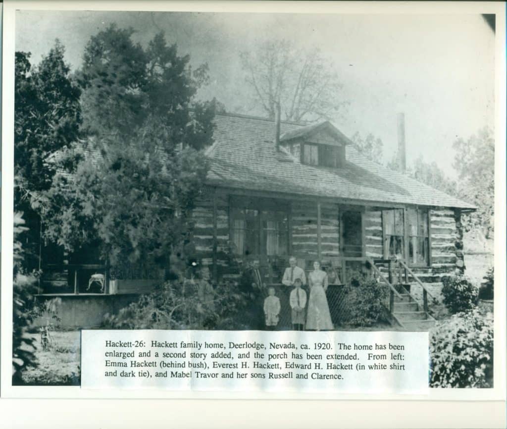 A black and white photo shows a family of five on the porch of a two-story log cabin.