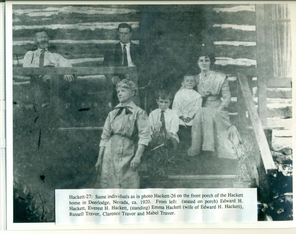 A black and white photo shows nine people posing on a rustic porch with a log wall.