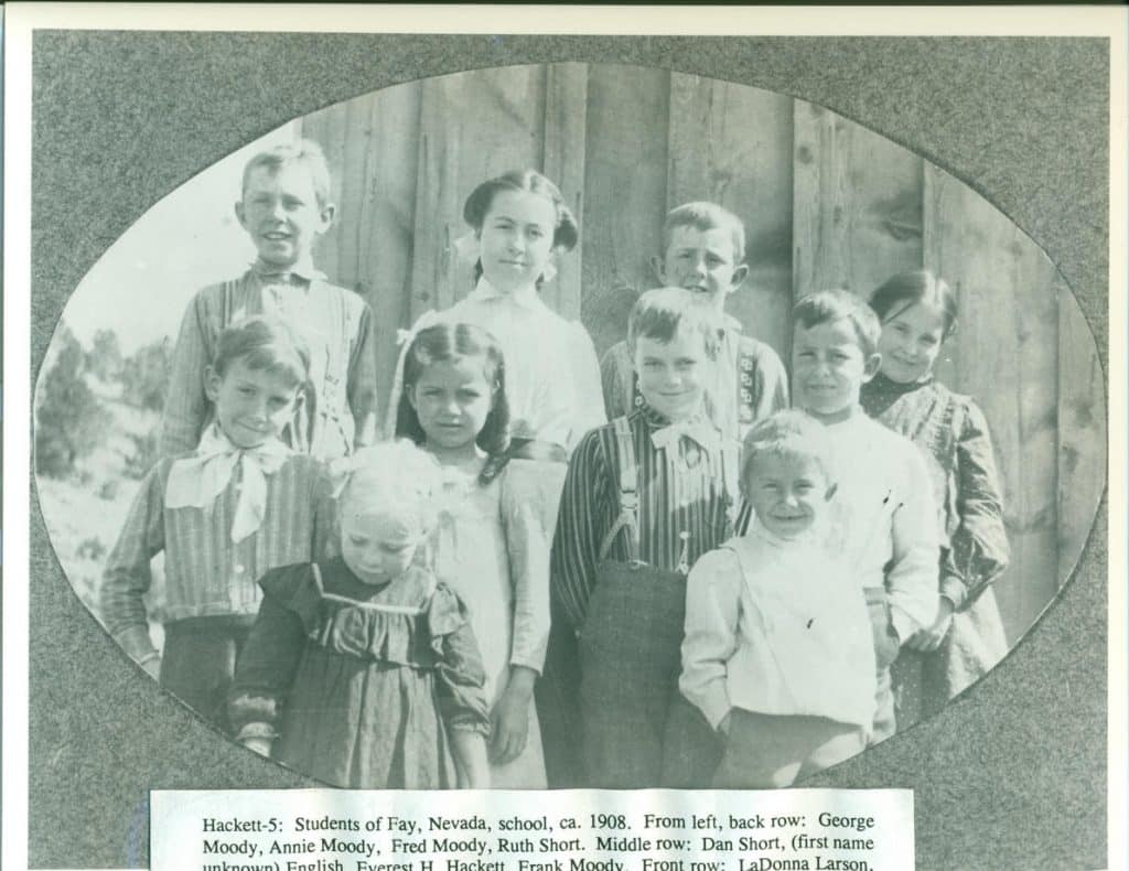 A black and white photo shows ten children, dressed in period clothing, posing in front of a wooden building.