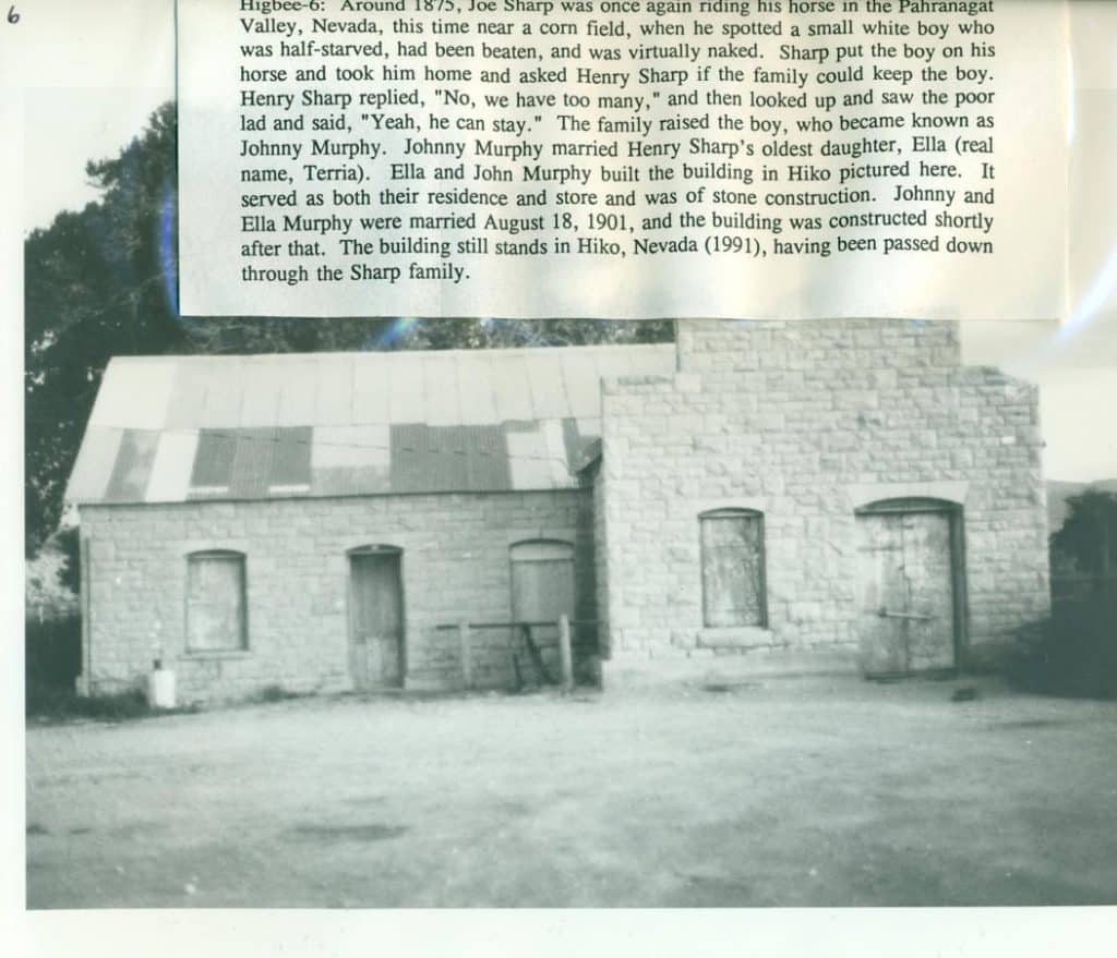 A black and white photo shows a stone building with a corrugated metal roof and three wooden doors.