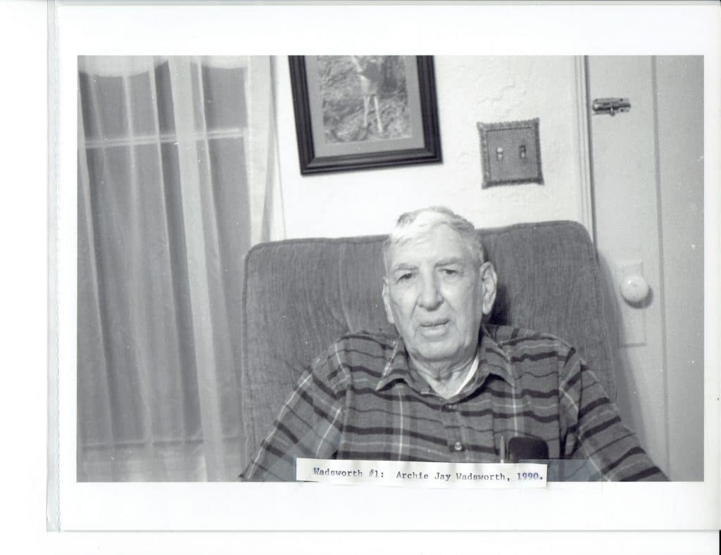 A black and white photo shows an older man with white hair in a plaid shirt sitting in a chair.