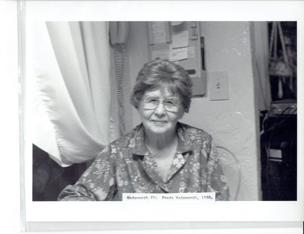 A smiling older woman with short hair and glasses wears a patterned shirt, sitting indoors.