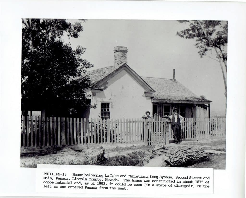 A black and white photo of a small house with a picket fence and two people standing behind it.