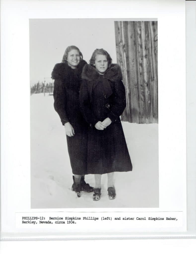 Two young women in dark coats stand in snow, with a wooden wall and fence in the background.