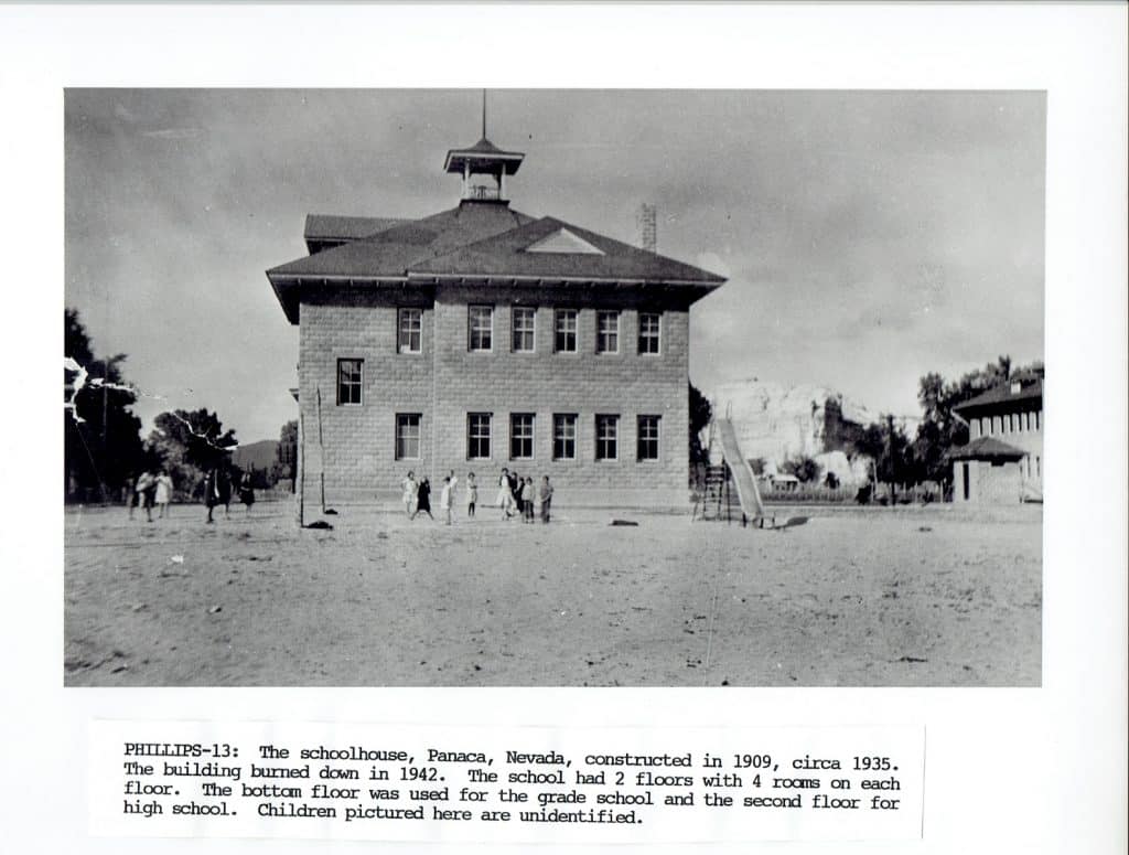 A black and white photo of a two-story brick schoolhouse with a bell tower and children playing outside.