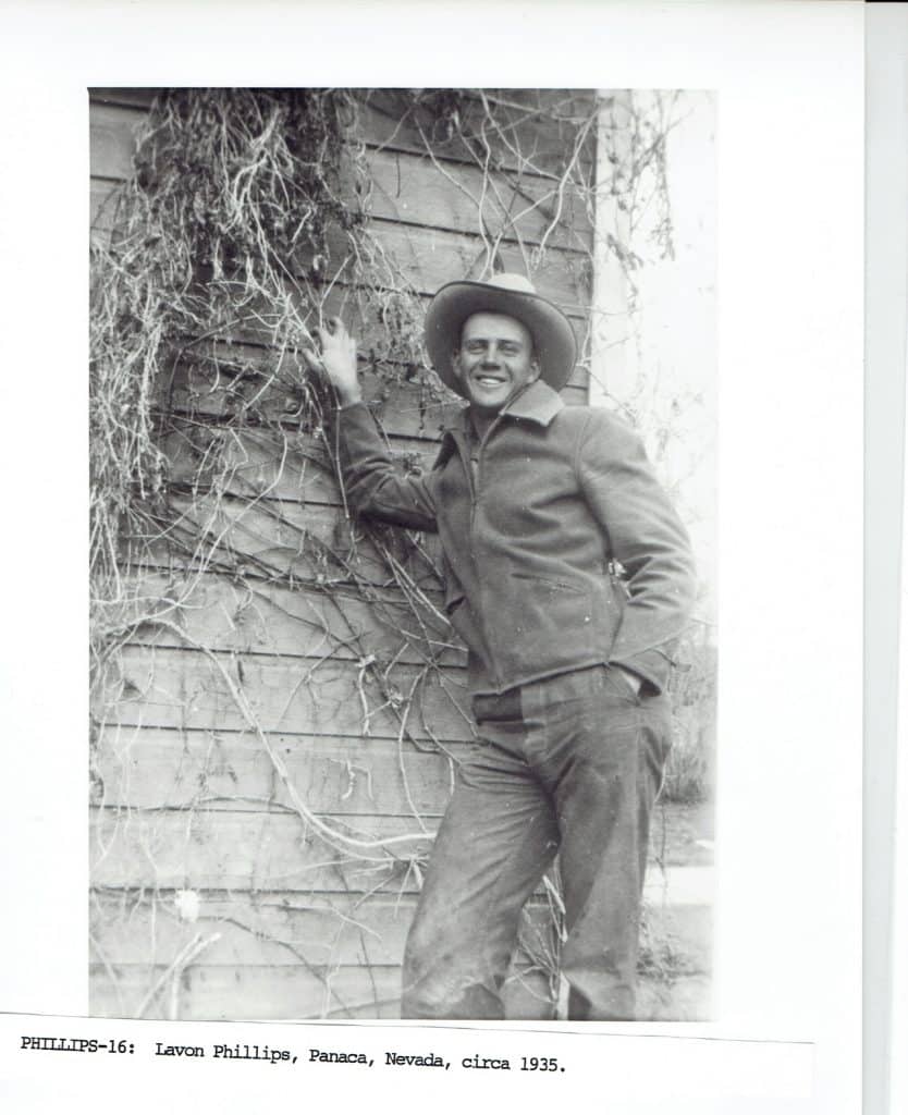 A smiling man in a cowboy hat, jacket, and jeans stands next to a wooden wall covered in vines.