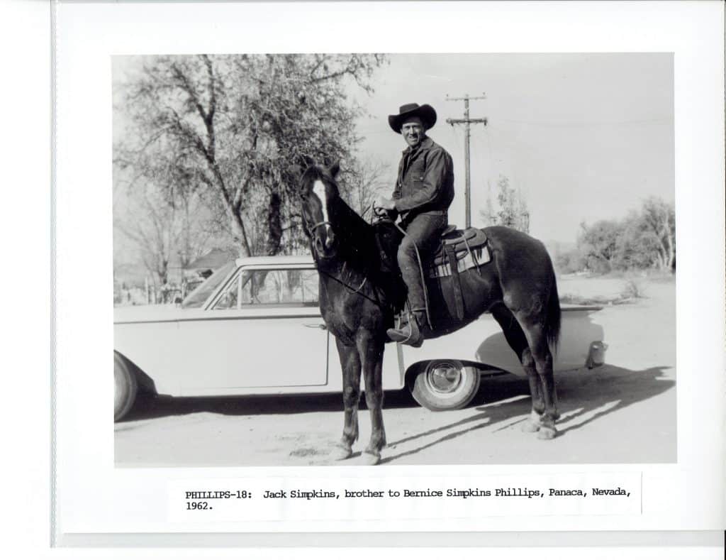 A smiling cowboy in a hat sits on a horse next to a white car on a dirt road.