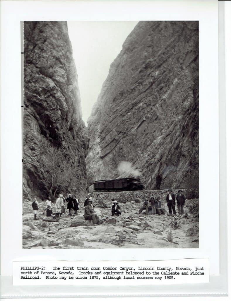 A black and white photo shows a steam train emerging from a canyon with people gathered in the foreground.