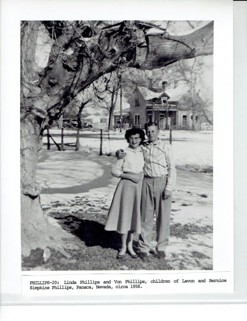 A black and white photo of a boy and girl standing arm-in-arm under a tree in front of a house.