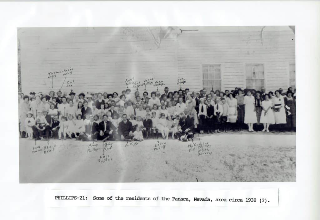 A large group of people, including men, women, and children, pose in front of a clapboard building.