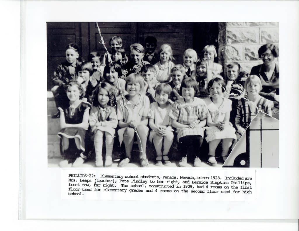 A black and white photo shows a group of smiling elementary school children and a teacher outdoors.
