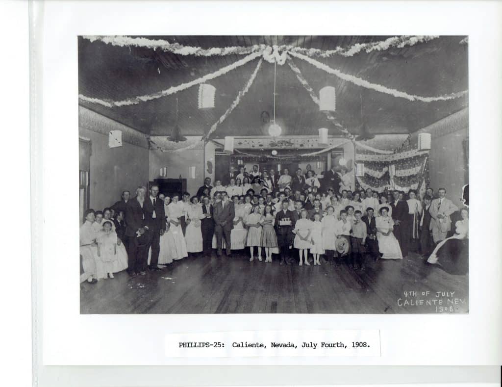 A large group of people, including many children, pose in a decorated hall for "4TH OF JULY CALIENTE NEV 1908."