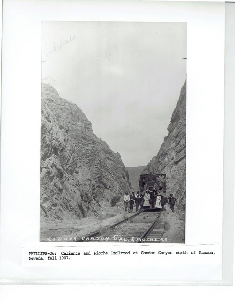 A black and white photo shows a steam train and seven people on tracks in a rocky canyon.