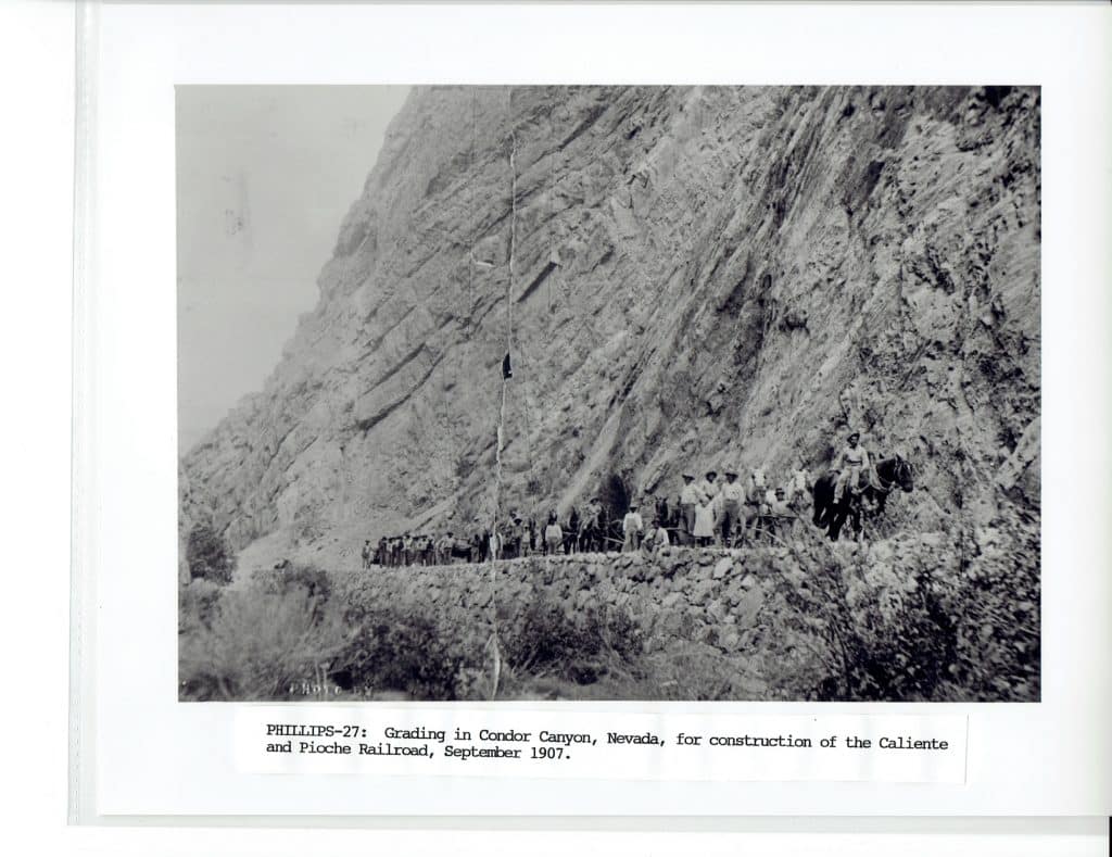 A black and white photo shows a large group of men and horses on a rocky ledge next to a steep mountain.